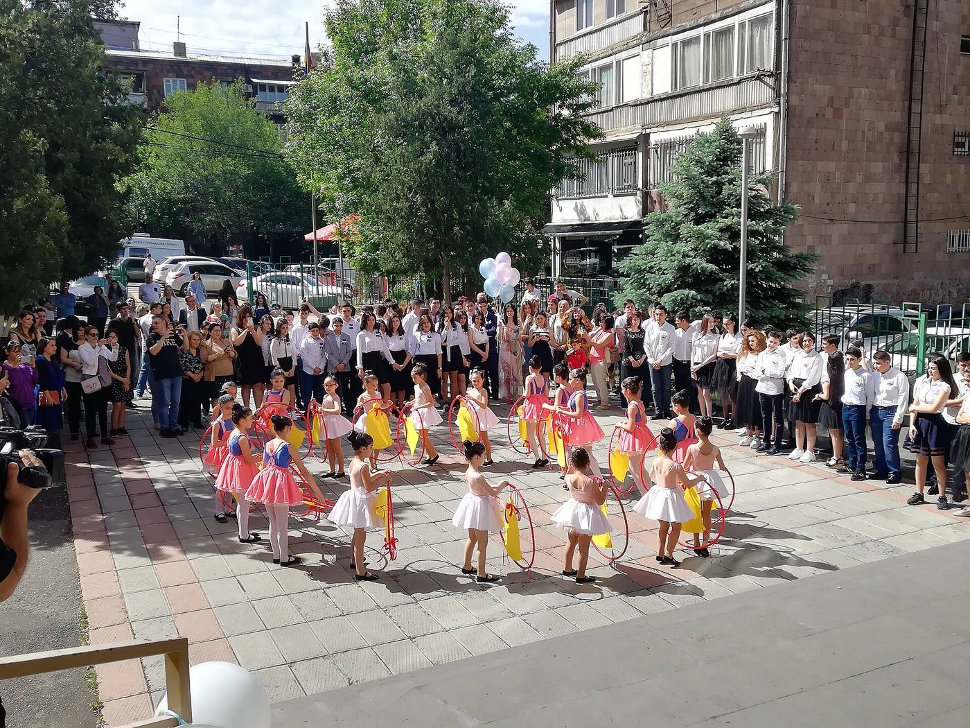 Девочки в пышных платьях с обручами выступают во дворе школы, вокруг стоят выпускники в белых рубашках и родители
