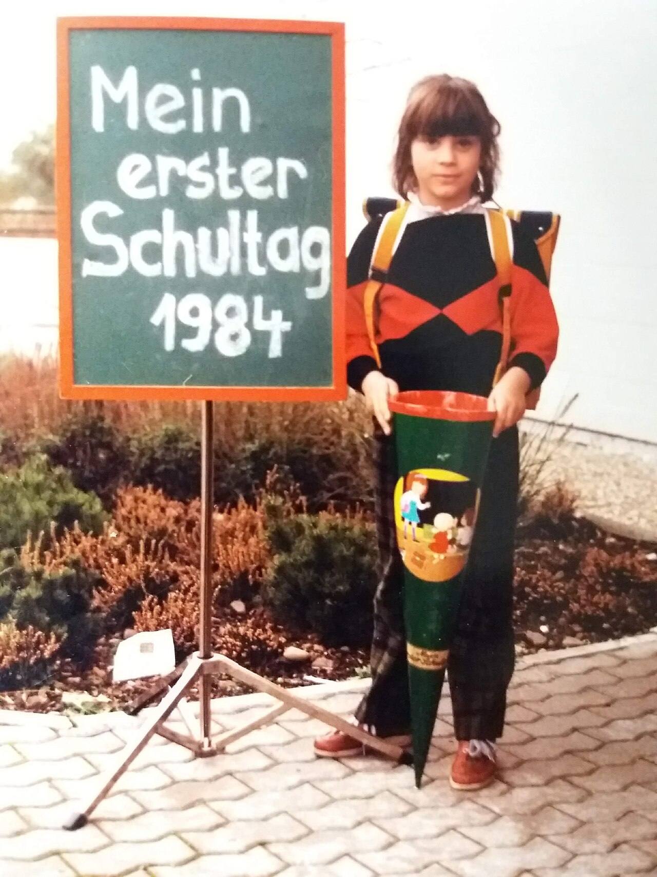 Девочка с ранцем и большим конусом Schultüte рядом с доской с надписью Mein erster Schultag 1984