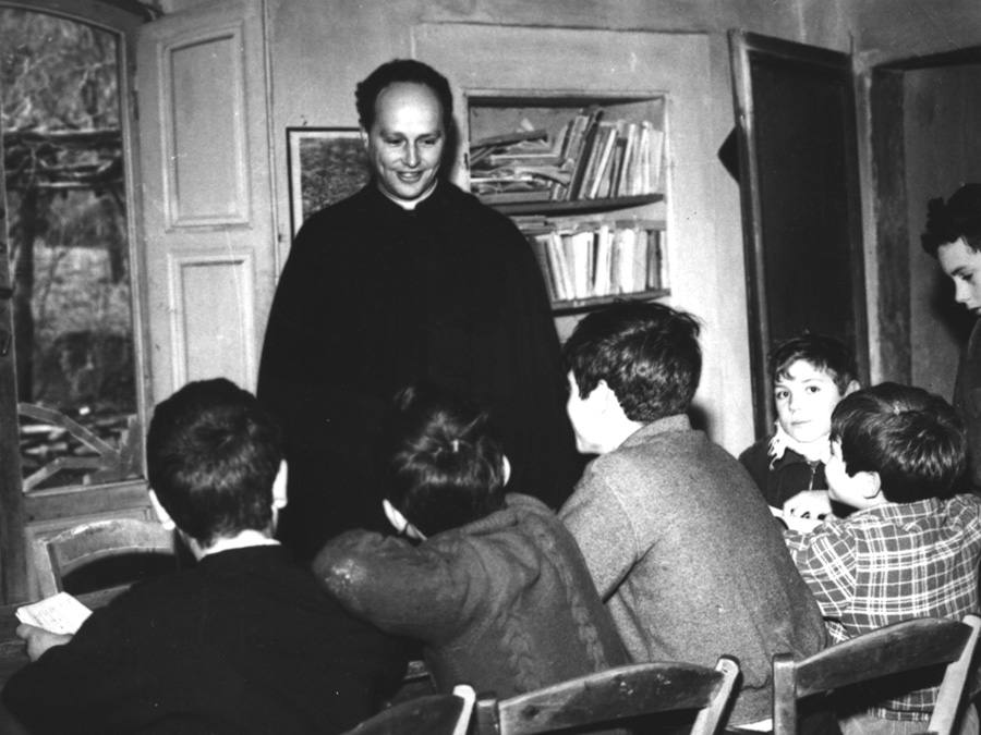 Священник стоит перед учениками в тесной комнате