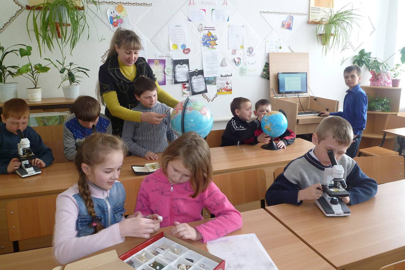 Учительница и ученики начальной школы с глобусами и микроскопами за партами